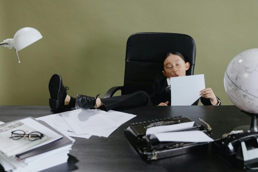 A child dressed in a suit sits in an office, surrounded by papers and a typewriter.